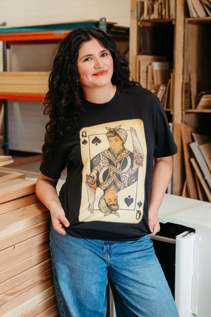 Woman leaning on lumber in a wood shop, wearing the black Queen of the Cut tee