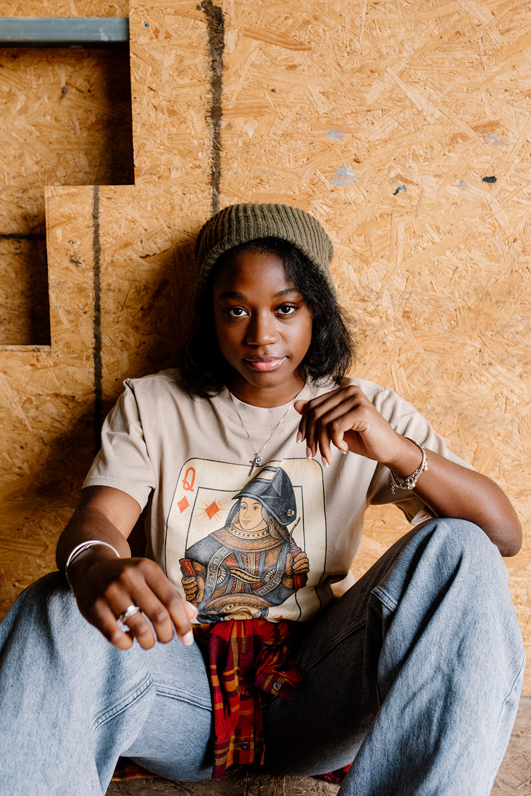 Woman sitting against a plywood wall wearing the ivory Queen of the Arc tee